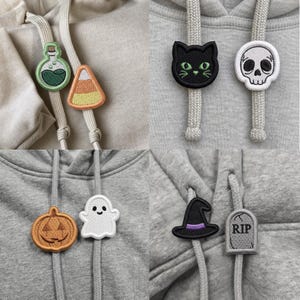 以下が含まれることがあります： 刺繍入りのドローストリングチャームが付いた4着のグレーのパーカー。デザインには、緑の薬瓶、キャンディコーン、黒猫、白い頭蓋骨、オレンジのカボチャ、白い幽霊、黒い魔女の帽子、RIPと書かれた灰色の墓石が含まれています。
