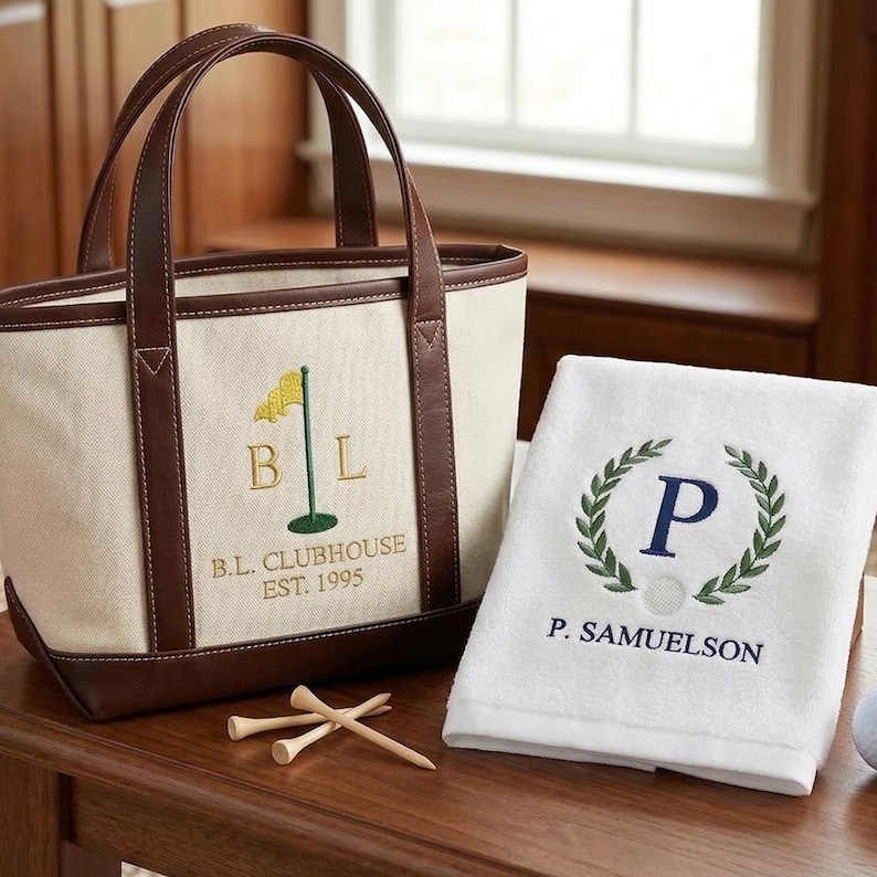 May include: A beige canvas tote bag with brown leather trim and handles, embroidered with a golf flag and the letters "BL". A white towel with a green laurel wreath and the letter "P" is next to the bag. Wooden golf tees are on the table.