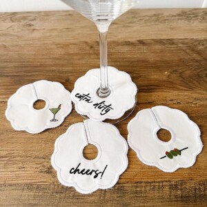 May include: White drink coasters with scalloped edges. One coaster has a martini illustration, another says "extra dirty", and a third says "cheers!". The fourth coaster has an olive illustration. A martini glass sits atop one coaster.