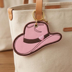 May include: Pink cowboy hat keychain with brown trim and a heart detail. The keychain is attached to a tan tote bag with a leather handle. A small gold star charm hangs from the keychain.