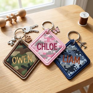 May include: Three diamond-shaped fabric keychains with metal rings. One features green camouflage with the name OWEN and a bear charm, one pink camouflage with CHLOE and a butterfly charm, and one blue camouflage with LIAM and an airplane charm.