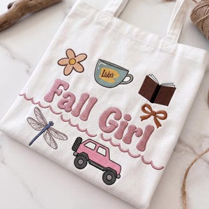 May include: White canvas tote bag with embroidered design. Features the text "Fall Girl" in pink, along with a flower, coffee cup, book, bow, dragonfly, and pink jeep. The bag has two handles.