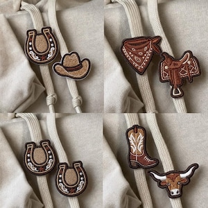 May include: A collection of embroidered patches featuring Western-themed designs including brown and tan horseshoes, a cowboy hat, a patterned bandana, a saddle, a leather boot with white stitching, and a longhorn steer head on a beige fabric.