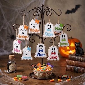 May include: A dark metal display stand holding ten white ghost-shaped ornaments, each featuring a unique name and a colourful bow or bowtie. The names include Evelyn, Ms. Sutton, Ozzie, Nics, Mia, Suncere, Boo, and Caleb, all hanging from twine.