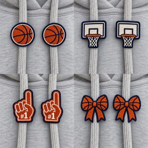 Op de afbeelding: Een set van acht geborduurde trekkoordbedels voor hoodies met basketbalthema. De collectie bevat oranje basketballen, witte basketbalringen, oranje nummer één schuimvingers en oranje strikken met een basketbalpatroon.