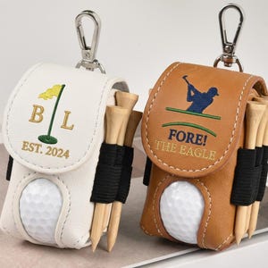 May include: Two golf ball pouches with carabiner clips. One is white with a green flag and the letters "B L" and "EST. 2024". The other is brown with a golfer graphic and the text "FORE! THE EAGLE". Each pouch holds golf tees and a golf ball.