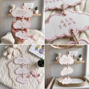 May include: A hanging nursery decoration featuring three stacked cloud-shaped plaques with pink embroidered edges. The plaques are connected by twine and display the text Maddox Turnbull, 6 lb 14 oz, and 19.10.25 in pink lettering.