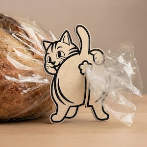 Op de afbeelding: Een beige stoffen broodclip in de vorm van een kat met zwarte geborduurde omlijningen. De kat is van achteren afgebeeld met de staart omhoog, en in het midden bevindt zich een kleine bloemvormige uitsparing. Het is bevestigd aan een plastic zak.