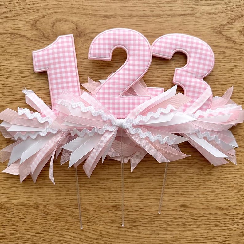 May include: Cake toppers with the numbers 1, 2, and 3. Each number is made of pink and white gingham fabric. The numbers are surrounded by pink and white ribbons and bows. Clear plastic sticks are attached to the bottom.