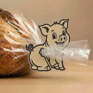 Op de afbeelding: Een beige, geborduurde broodclip in de vorm van een varken met zwarte omlijningen en een krulstaart. Het varken is bevestigd aan een doorzichtige plastic zak met brood, waarbij het plastic door een uitsparing in het midden is gehaald.