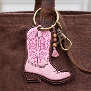 May include: Pink cowboy boot keychain with embroidered details and a brown border. The keychain is attached to a bronze-colored clasp and a ring, with wooden beads and a pink tassel. The background is a brown canvas bag.