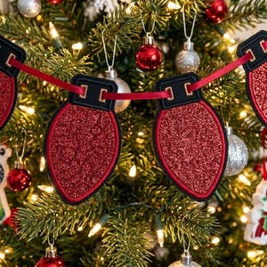 May include: A festive garland featuring multiple red glittery light bulb-shaped ornaments connected by a thin red ribbon. Each bulb has a black stitched top and a red border, creating a decorative string of holiday lights hanging on a tree.