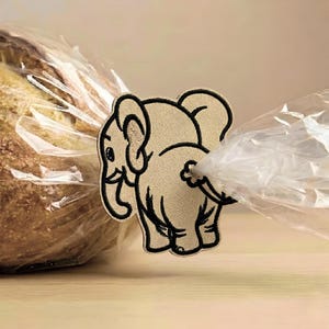 Op de afbeelding: Een beige geborduurde olifantenpatch met zwarte omlijningen, die fungeert als broodzakclip. De olifant is afgebeeld in zijaanzicht, met de slurf naar links gericht en de staart gepositioneerd om de gedraaide doorzichtige plastic zak vast te houden.