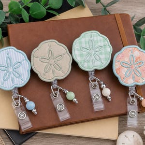 May include: Four retractable badge reels featuring embroidered sand dollar designs in light blue striped, solid beige, light green gingham, and peach floral fabric patterns, each with a clear plastic strap and a small decorative bead charm.