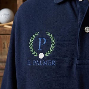 May include: Navy blue polo shirt with embroidered design. The design includes a green laurel wreath, a blue letter "P", and the name "S. Palmer" in blue. A white golf ball is also visible.