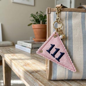 May include: A pink pennant-shaped keychain with the number "111" in navy blue. The keychain is attached to a striped tote bag with a gold-colored clasp and small charms, including a pearl and a star.
