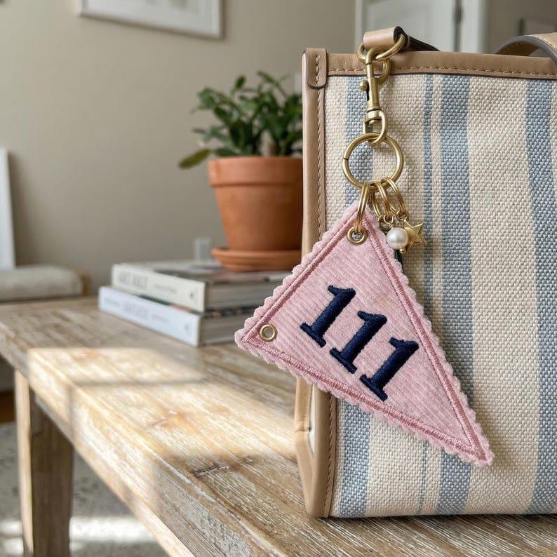 May include: A pink pennant-shaped keychain with the number "111" in navy blue. The keychain is attached to a striped tote bag with a gold-colored clasp and small charms, including a pearl and a star.