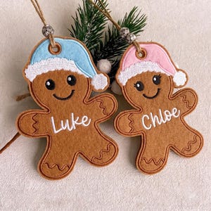 Puede incluir: Dos adornos de fieltro con forma de hombre de jengibre y detalles bordados. Uno lleva un gorro azul claro y tiene bordado el nombre Luke. El otro lleva un gorro rosa claro y tiene bordado el nombre Chloe. Ambos tienen cordeles con cuentas plateadas.