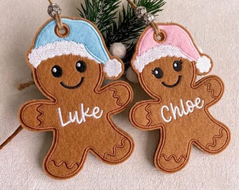 May include: Two felt gingerbread person ornaments with embroidered details. One wears a light blue hat and is embroidered with the name Luke. The other wears a light pink hat and is embroidered with the name Chloe. Both have twine hangers with silver beads.