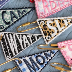 May include: Embroidered pennant keychains in various patterns and colors, including leopard print, gingham, and floral designs. Each keychain features a gold-tone clasp and embroidered lettering spelling out names and words like "MOM" and "WYATT".