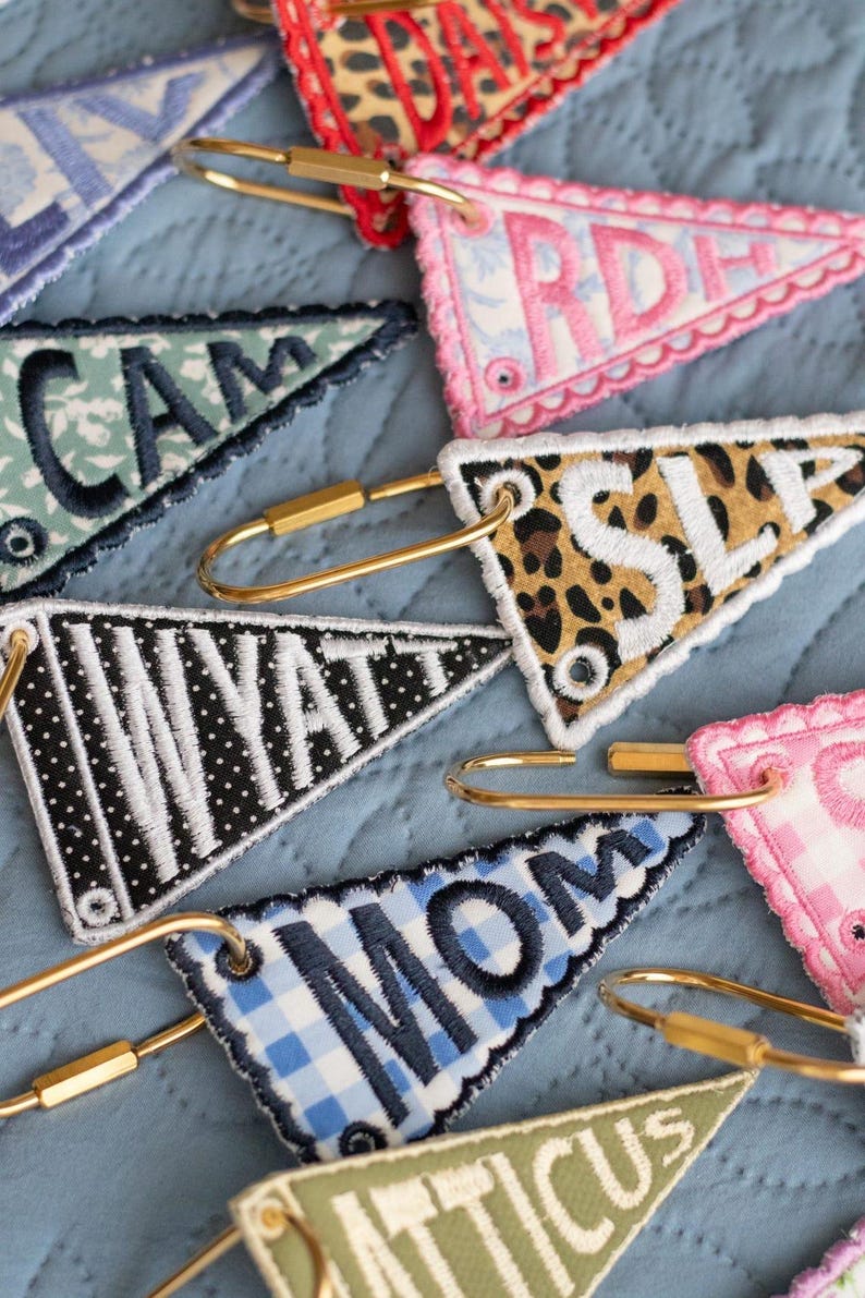 May include: Embroidered pennant keychains in various patterns and colors, including leopard print, gingham, and floral designs. Each keychain features a gold-tone clasp and embroidered lettering spelling out names and words like "MOM" and "WYATT".