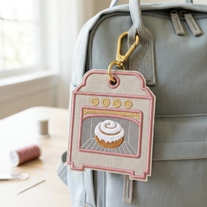 Puede incluir: Una mochila gris claro con una etiqueta bordada colgante con forma de horno. La etiqueta presenta un borde rosa, cuatro perillas circulares amarillas y un gráfico central de un rollo de canela marrón y blanco horneándose en el interior.