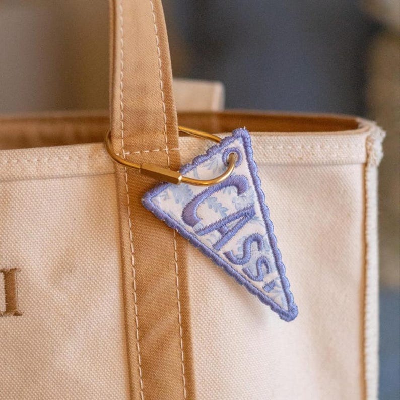 K&ouml;nnte beinhalten: Eine beigefarbene Canvas-Tragetasche mit einem dreieckigen Stoff-Schl&uuml;sselanh&auml;nger an einem goldfarbenen Metallring. Der Anh&auml;nger hat ein hellblaues Blumenmuster, den Namen CASSI in violetter Stickerei und einen passenden violetten Rand.