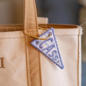 May include: A cream-colored tote bag with tan handles and a small, triangular, embroidered tag. The tag is light blue with a floral pattern and the name "CASS" in a darker blue. The tag is attached to the bag with a gold-colored clasp.