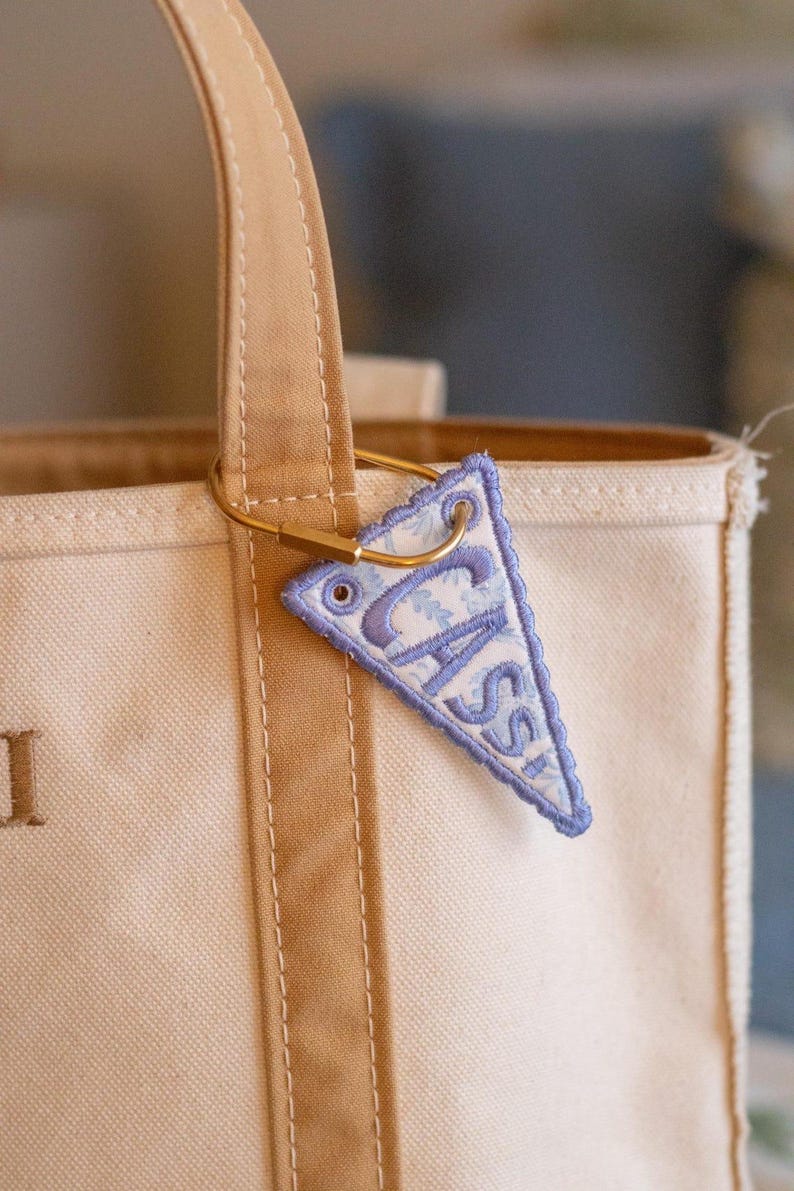 May include: A cream-colored tote bag with tan handles and a small, triangular, embroidered tag. The tag is light blue with a floral pattern and the name "CASS" in a darker blue. The tag is attached to the bag with a gold-colored clasp.