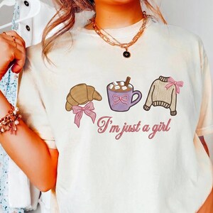 May include: Cream-colored t-shirt with embroidered graphics of a croissant, a mug of hot chocolate, and a sweater, each adorned with pink bows. The phrase "I'm just a girl" is written in cursive below the graphics.