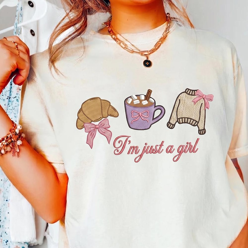 May include: Cream-colored t-shirt with embroidered graphics of a croissant, a mug of hot chocolate, and a sweater, each adorned with pink bows. The phrase "I'm just a girl" is written in cursive below the graphics.