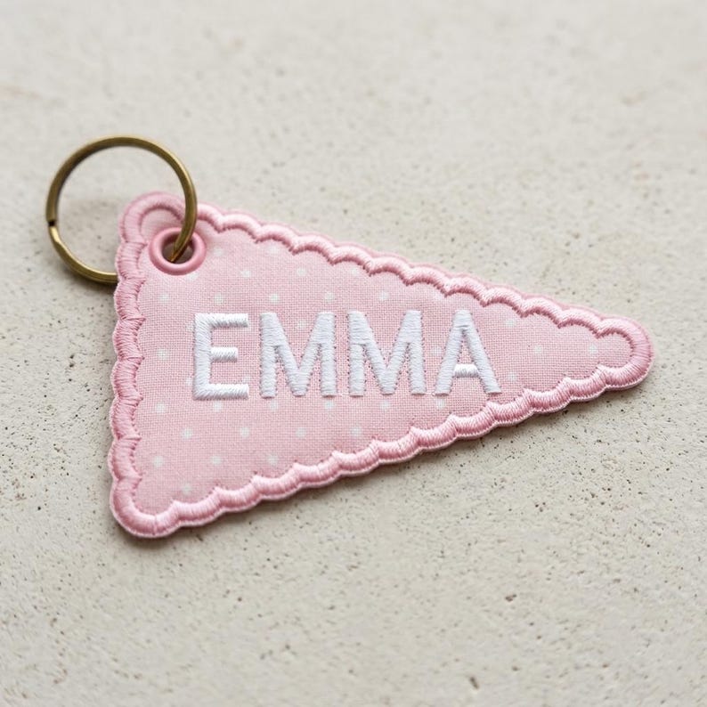 K&ouml;nnte beinhalten: Ein dreieckiger Schl&uuml;sselanh&auml;nger aus Stoff in Hellrosa mit wei&szlig;em Punktemuster und gewelltem Rand. Der Name EMMA ist in wei&szlig;en Gro&szlig;buchstaben in die Mitte gestickt. Ein runder, bronzefarbener Metallring ist an der oberen Ecke befestigt.
