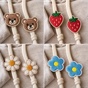 Op de afbeelding: Een verzameling van vier paar geborduurde patches bevestigd aan dikke, crèmekleurige gevlochten koorden. De ontwerpen omvatten bruine berengezichten, rode aardbeien met groene blaadjes, witte madeliefjes en blauwe bloemen met een geel hart.
