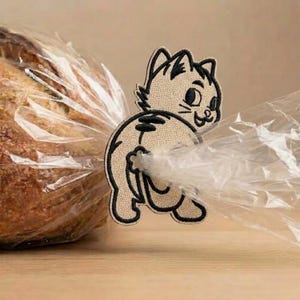 Pode incluir: Um clipe de tecido bege para saco de pão em forma de gato de desenho animado com contornos e detalhes bordados a preto. A figura do gato está posicionada para segurar um saco de plástico transparente que contém um pão.
