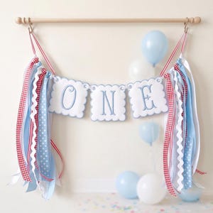 May include: A festive banner spelling "ONE" in blue script on white scalloped panels, adorned with red and blue ribbons and balloons. The banner is suspended from a wooden rod, perfect for a first birthday celebration.
