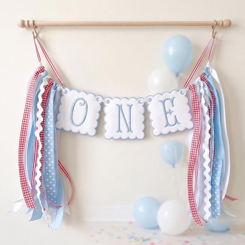 May include: A festive banner spelling "ONE" in blue script on white scalloped panels, adorned with red and blue ribbons and balloons. The banner is suspended from a wooden rod, perfect for a first birthday celebration.