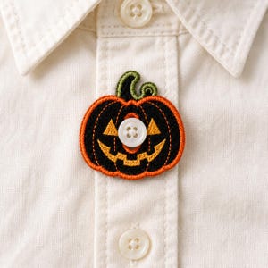 Puede incluir: Una camisa blanca de botones con un parche bordado de calabaza de Halloween. El parche es negro con borde naranja, tallo verde, ojos triangulares amarillos y una boca dentada. Un botón blanco de cuatro agujeros está centrado en el parche.