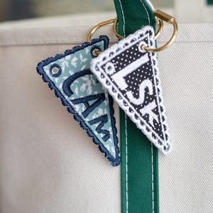 May include: Two personalized pennant keychains with gold rings hang from a green and white tote bag strap. One pennant is light blue with white floral print and the letters "CAM". The other is black and white polka dots with the letters "LSK".