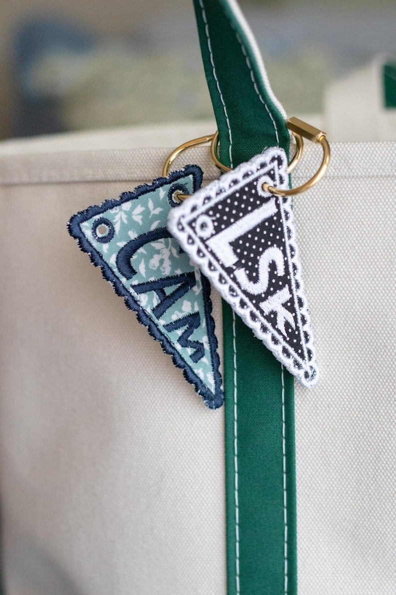May include: Two personalized pennant keychains with gold rings hang from a green and white tote bag strap. One pennant is light blue with white floral print and the letters "CAM". The other is black and white polka dots with the letters "LSK".