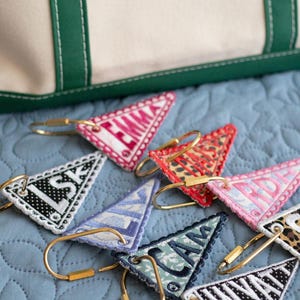 May include: A collection of personalized triangular keychains with gold-colored hardware, each embroidered with a different name or initials. The keychains feature various patterns and colors, including pink, red, and black, and are arranged on a blue quilted surface.