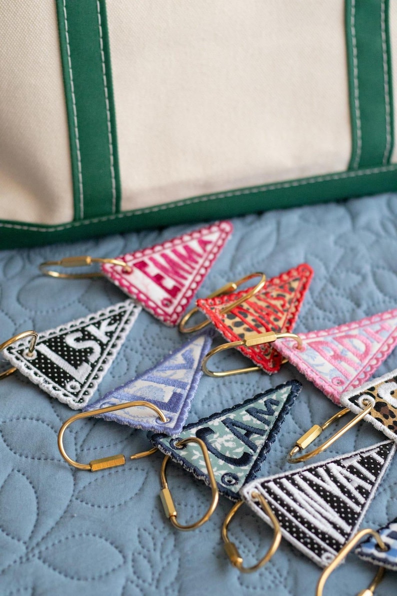 May include: A collection of personalized triangular keychains with gold-colored hardware, each embroidered with a different name or initials. The keychains feature various patterns and colors, including pink, red, and black, and are arranged on a blue quilted surface.