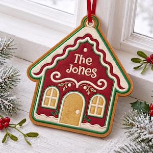 May include: A house-shaped Christmas ornament featuring a deep red fabric center with the embroidered text "The Jones". The design includes a gold door, two windows, white icing-style trim, green borders, and a red ribbon loop for hanging.