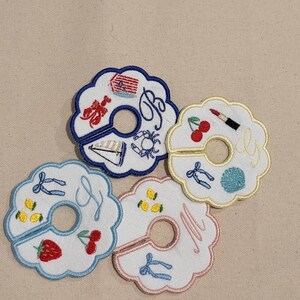 May include: Embroidered white fabric coasters with scalloped edges. Each coaster features a unique design, including a lobster, sailboat, cherries, and lemons. The coasters have a hole in the center and embroidered letters.