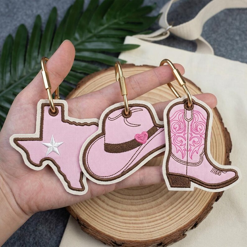 May include: Three pink and brown embroidered keychains with gold-colored clasps. The keychains are shaped like the state of Texas, a cowboy hat with a heart, and a cowboy boot. The Texas keychain has a white star.