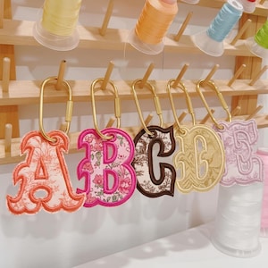 Op de afbeelding: Vijf geborduurde letter-sleutelhangers met de letters A, B, C, D en E, elk bevestigd aan een goudkleurige ovale karabijnhaak. De letters tonen verschillende stofpatronen, waaronder bloemen, toile en effen texturen met gestikte randen.