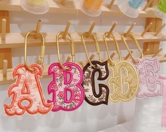 May include: Five embroidered letter keychains featuring the letters A, B, C, D, and E, each attached to a gold-toned oval carabiner. The letters display various fabric patterns including floral, toile, and solid textures with stitched borders.
