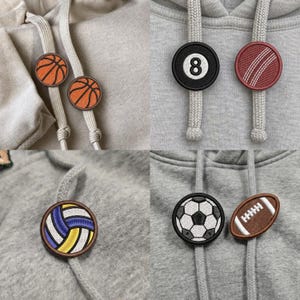 May include: A collection of circular and oval embroidered patches featuring sports themes attached to gray hoodie drawstrings. The designs include a basketball, an eight ball, a cricket ball, a volleyball, a soccer ball, and an American football.