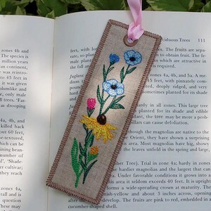 以下が含まれることがあります： 青、ピンク、黄色の花柄が刺繍されたリネンのブックマーク。ブックマークにはピンクのリボンと茶色の縁取りがあります。ブックマークの長さは約18cmです。
