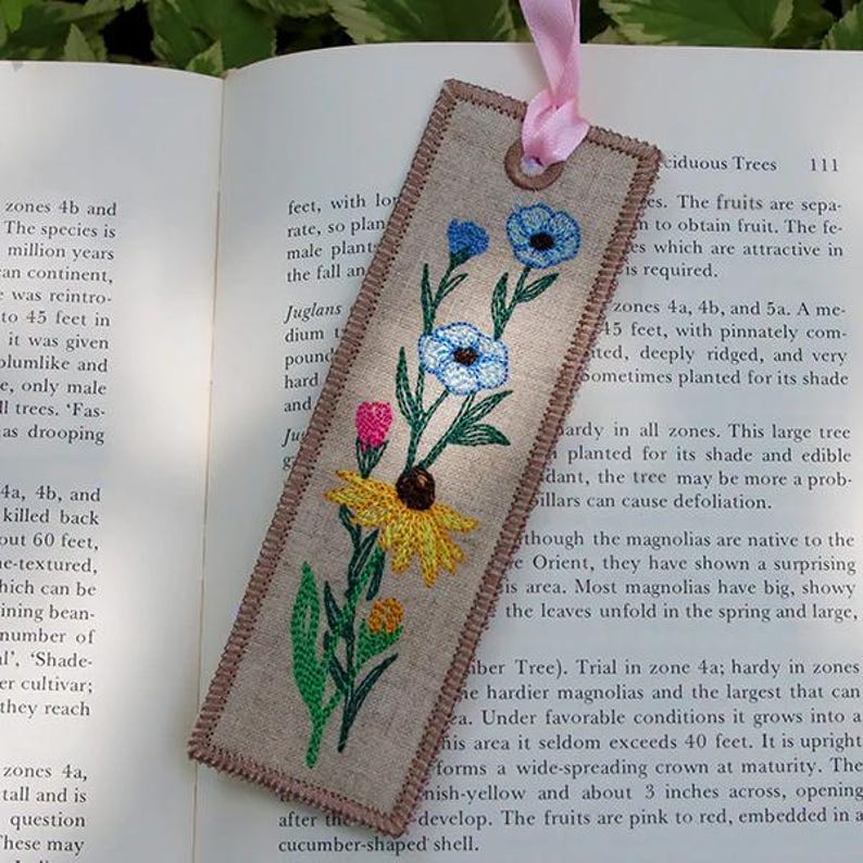 以下が含まれることがあります： 青、ピンク、黄色の花柄が刺繍されたリネンのブックマーク。ブックマークにはピンクのリボンと茶色の縁取りがあります。ブックマークの長さは約18cmです。