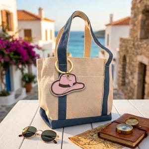 May include: A beige tote bag with navy blue accents and handles, featuring a pink cowboy hat keychain. The bag is on a white table with sunglasses, a compass, and a map. The background shows a coastal town.